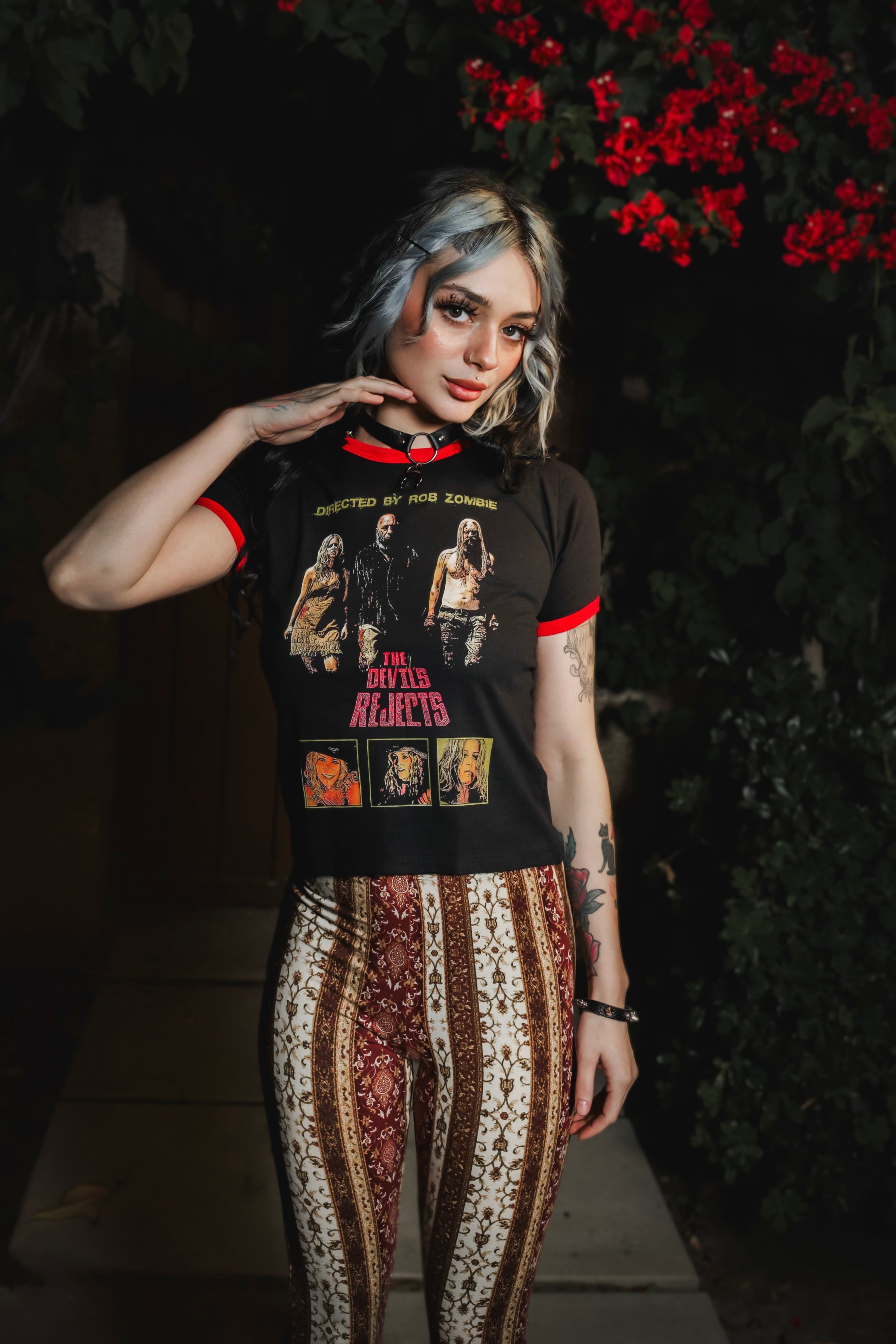 Person wearing a black t-shirt with graphic designs and patterned pants standing against a dark background with red flowers.