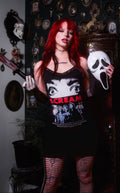 Person wearing a 'Scream' tank top holding a white mask in a darkly decorated room.
