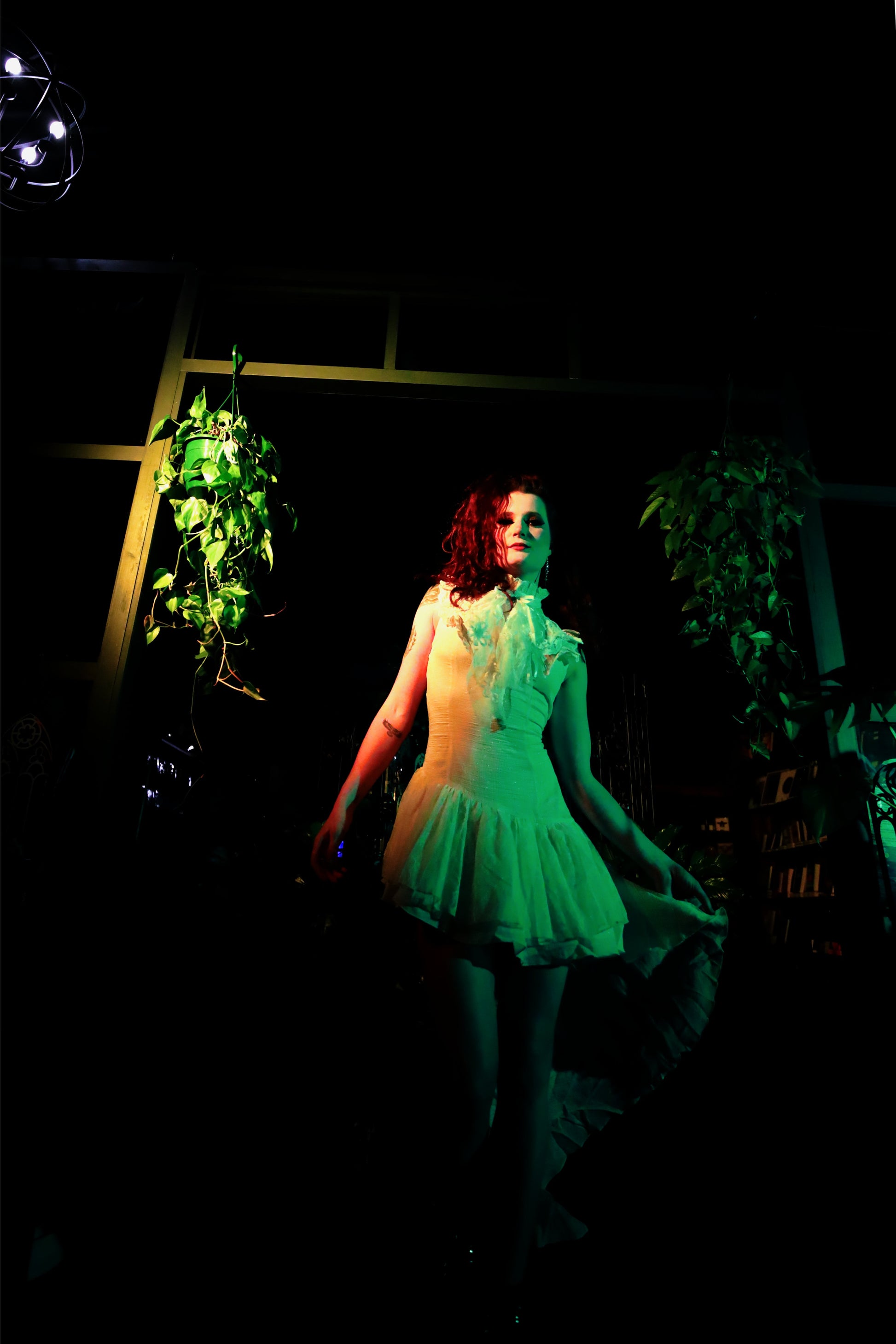 Person in a white asymmetrical dress standing in a dark room with plants with green and orange lighting.