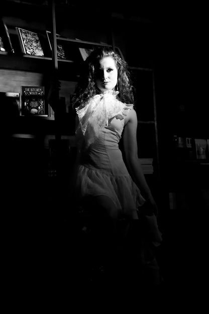 Black and white photo of a person in a vintage dress sitting in a dimly lit room in front of old library shelves and a ladder.
