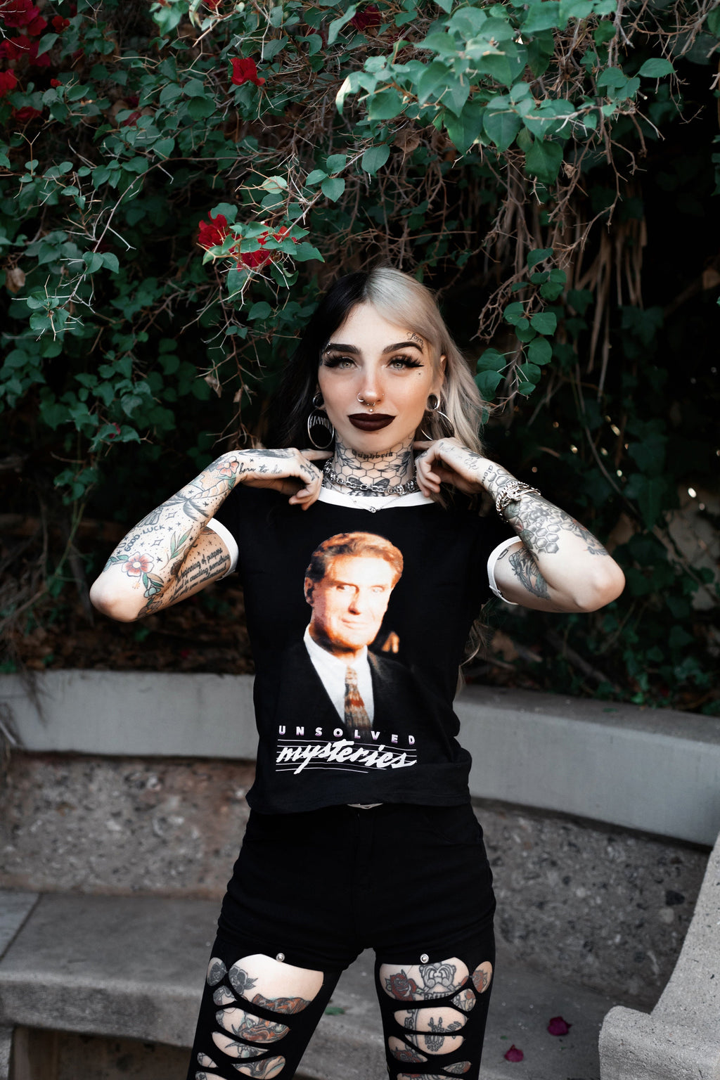 Person wearing a black t-shirt that has Robert Stack's face and says 'Unsolved Mysteries' with a graphic design, standing outdoors with greenery in the background.